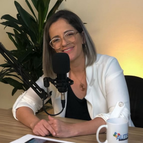 Foto da palestrante Sabrina Barcelos, sentada à mesa de uma estação rádio, falando ao microfone. Sabrina veste uma blusa branca de meia manga, usa óculos e tem cabelos claros na altura do ombro. Ao fundo há uma planta decorativa e a parede de cor alaranjada. Sobre a mesa estão um smartphone e uma caneca com a palavra autismo.